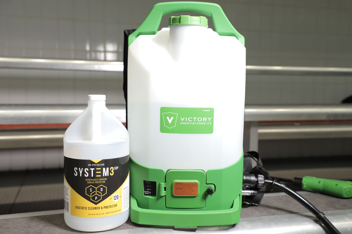 The Layer 1 surface product in its ready-to-use gallon beside a backpack electrostatic sprayer, on facility bleachers.
